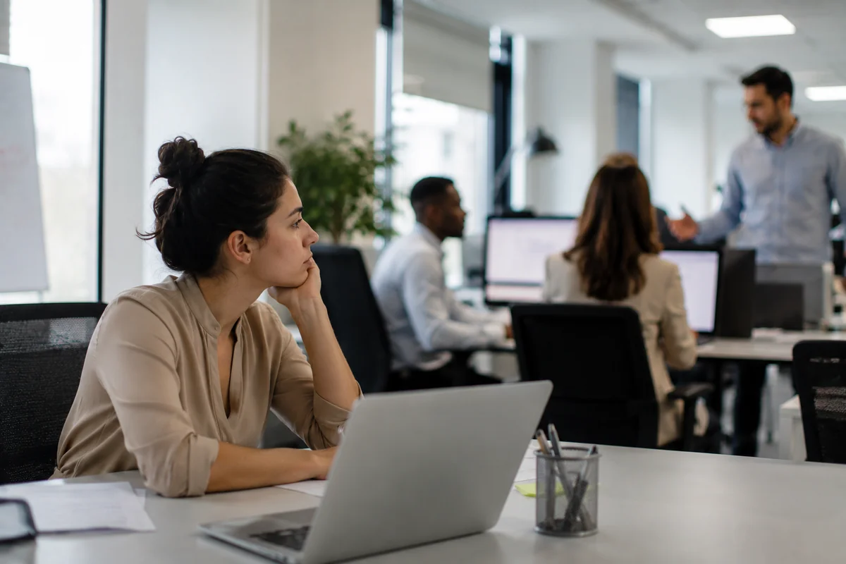A young woman in a beige blouse sitting at her office desk, resting her chin on her hand and gazing off pensively, while colleagues work and talk together in the background, conveying feeling invisible and overlooked at work