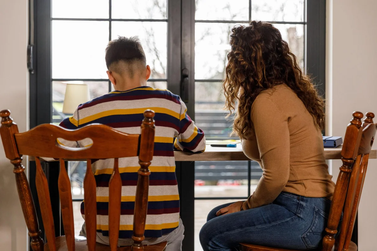 A parent sitting beside their child at a table by a window while the child works on schoolwork, offering quiet support, illustrating how parents can help a child cope with exam stress