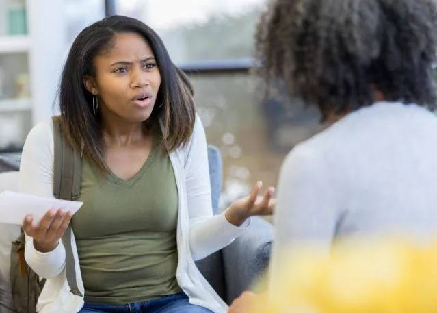 An upset teenage girl gesturing and speaking intensely while holding a piece of paper, sitting across from an adult woman seen from behind